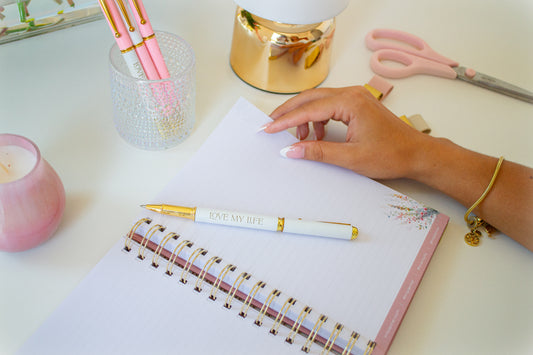 Person holding a pen over an open notebook with decorative elements on a desk.