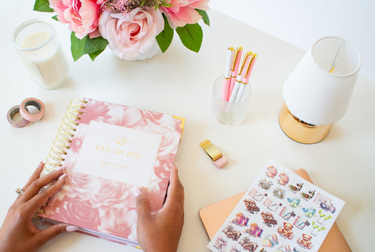 Person holding a pink planner with decorative elements on a desk.