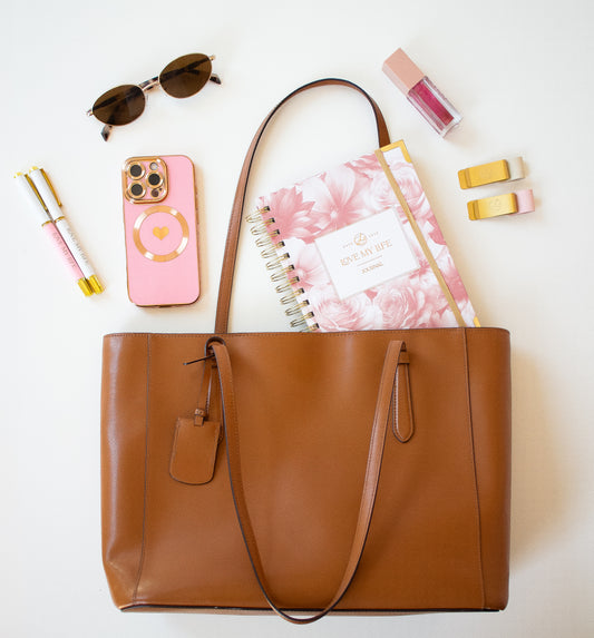 Brown tote bag with pink floral notebook, phone, sunglasses, and other items on a white background