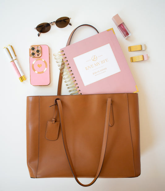 Brown tote bag with pink notebook, phone, sunglasses, and other items on a white background