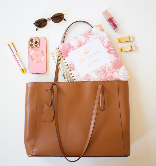 Brown tote bag with floral notebook, phone, sunglasses, and other items on a white background