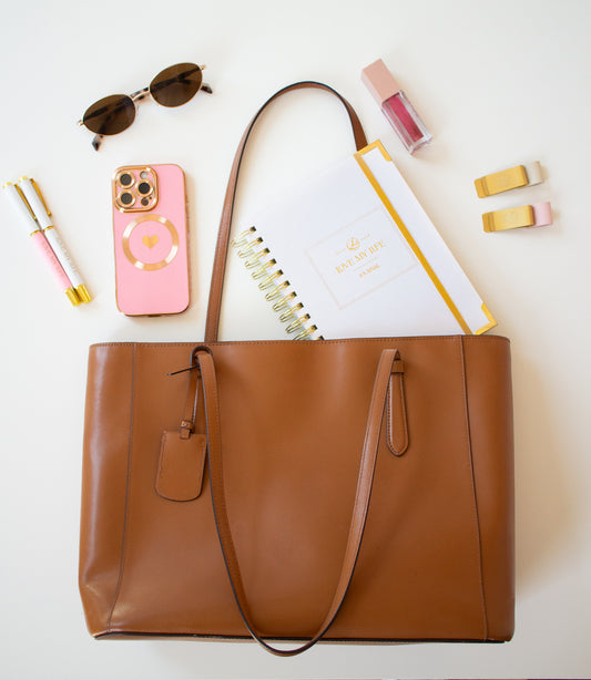 Brown tote bag with phone, sunglasses, notebook, and stationery items on a white background