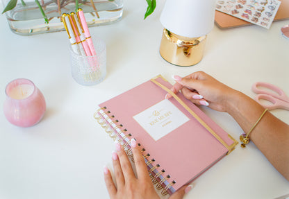 Person holding a pink notebook with a gold spiral on a white surface, surrounded by stationery items.