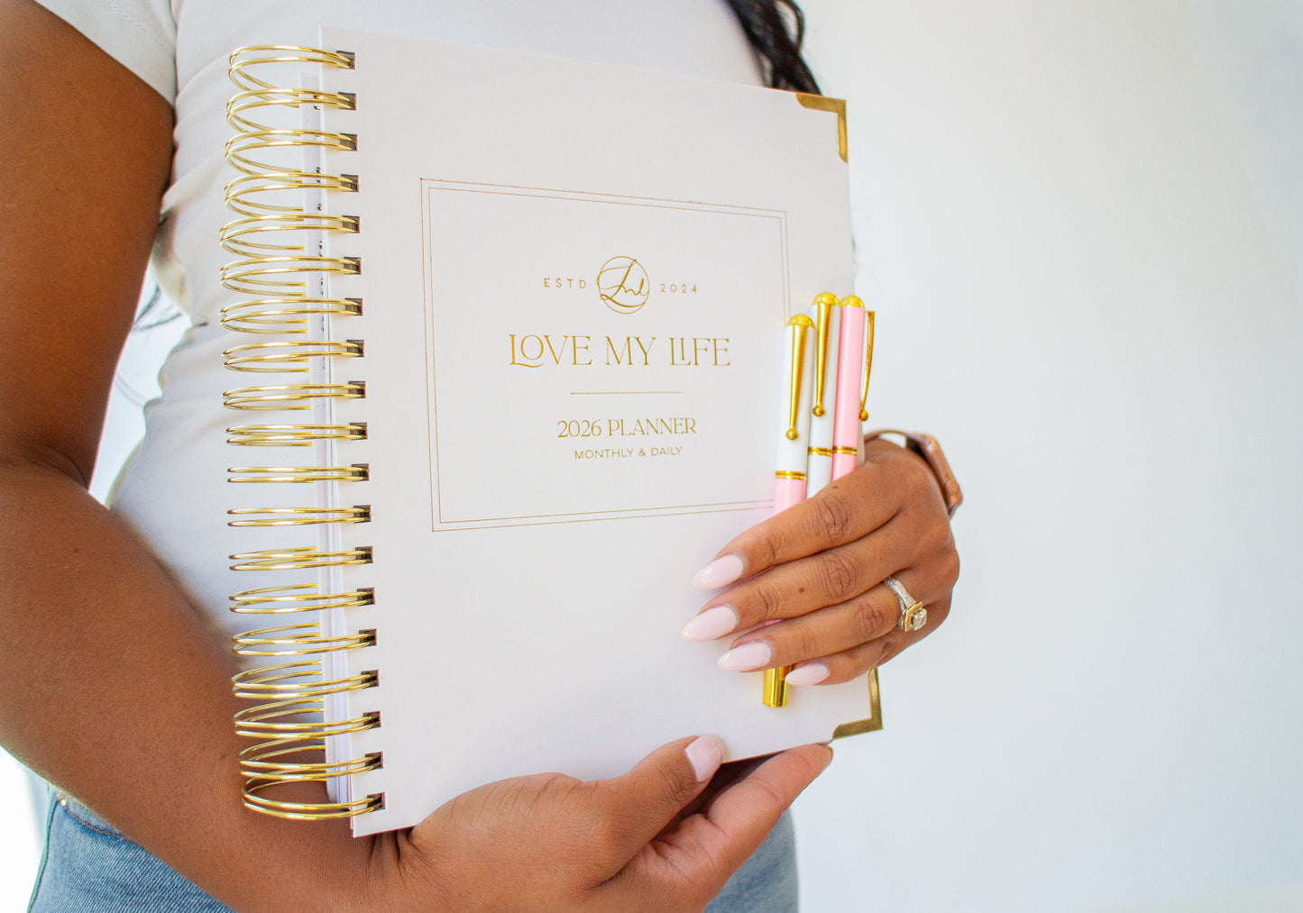 Person holding a planner with gold spiral binding and pink pens on a white background