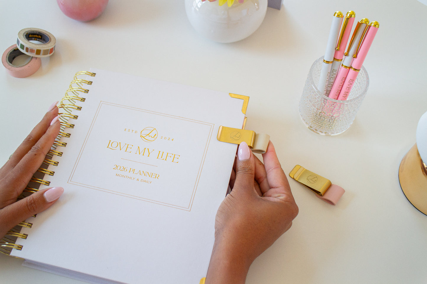 Person holding a gold clip over a notebook with stationery items on a desk.