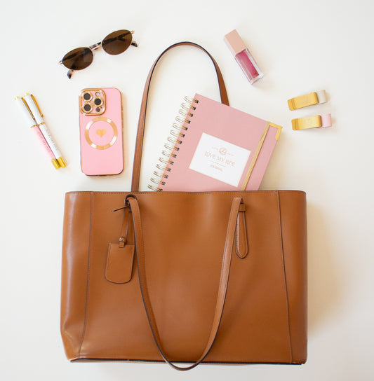 Brown tote bag with pink phone, notebook, sunglasses, and hair clips on a white background